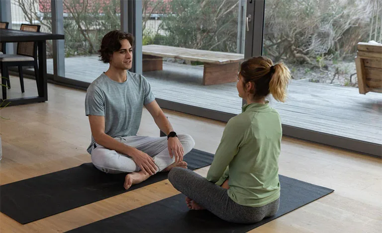 Two calm people sit cross-legged on mats by a sunlit window, relaxed and at ease together